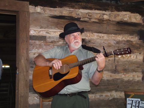 1966 Ford Ranchero - A man with a white beard and a black hat plays an acoustic guitar indoors.