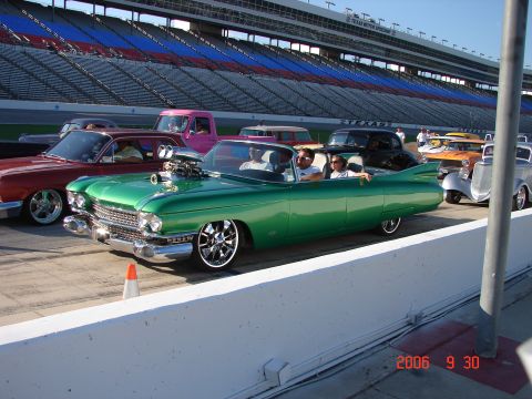 1987 Ford Bronco - Several custom cars are lined up at a racetrack, with grandstands in the background.