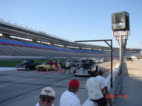 1987 Ford Bronco - Cars and trucks gathered at a racetrack with stadium seating in the background.