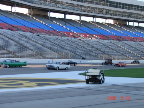 1987 Ford Bronco - A collection of classic cars are on display at a racetrack, with grandstands in the background.