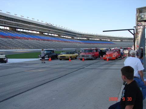 1987 Ford Bronco - A variety of vintage vehicles are lined up on a paved surface, with a large stadium in the background.
