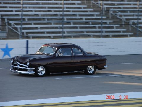 1987 Ford Bronco - 1950 Ford Custom Coupe on a track with stadium seating in the background.