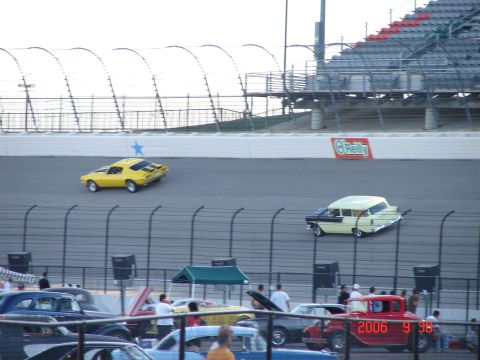 1987 Ford Bronco - Two classic cars race on a track, with spectators in the stands and other vehicles parked below.