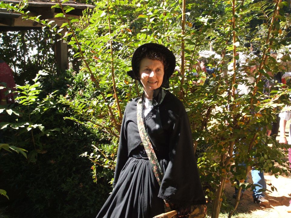 Woman in black historical dress and hat stands in front of green foliage