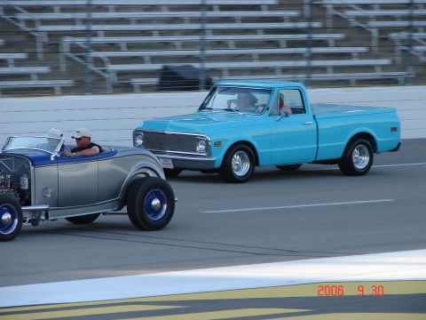 1987 Ford Bronco - A blue pickup truck and a silver roadster race on a track, with stadium seating in the background.