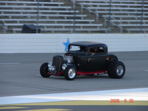 1987 Ford Bronco - A black hot rod coupe with a prominent engine is seen on a race track with grandstands in the background.