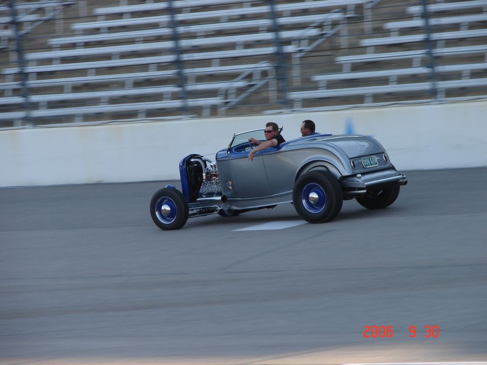 Silver and blue hot rod with two occupants driving on a track