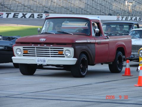 1987 Ford Bronco - 1965 Ford F-100 pickup truck parked at a racetrack with other vehicles.