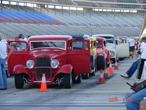 1987 Ford Bronco - Several classic hot rod trucks are lined up on a track, with stadium seating in the background.