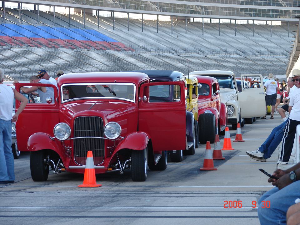 Line of hot rod trucks, red one in front with doors open, at a race track