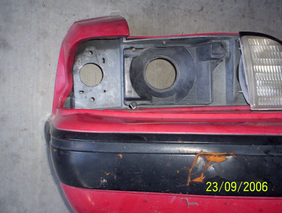 Red car bumper with black trim and a damaged headlight assembly, showing rust spots