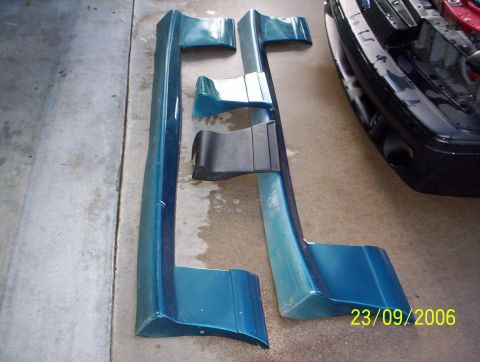 1990 Ford Mustang - Teal car side skirts and bumper trim pieces are laid out on a concrete surface next to a black car bumper.