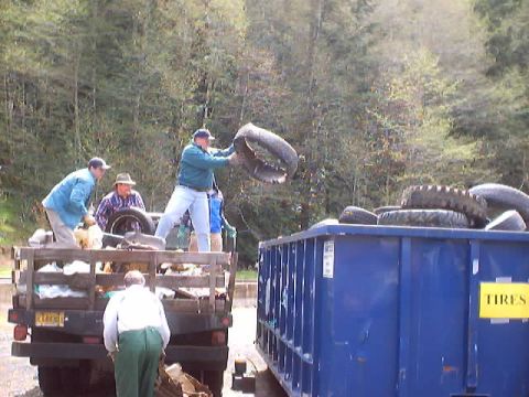 1976 Ford F-100 - People are disposing of old tires by loading them into a large blue dumpster.