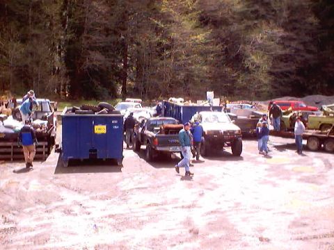 1976 Ford F-100 - A scene of multiple trucks and people in a salvage yard, with large blue dumpsters present.