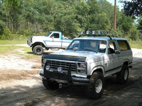1986 Ford Bronco - Two modified Ford trucks parked outdoors on a dirt track with trees in the background.