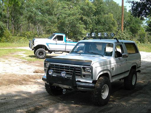 White 1980s Ford Bronco with roof lights and a white Ford pickup truck with blue trim in the background