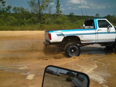 1986 Ford Bronco - A lifted pickup truck navigates through a muddy trail, with another vehicle visible in the side mirror.