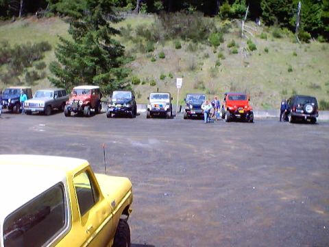 1976 Ford F-100 - A group of off-road vehicles are parked in a lot, with a wooded hillside behind them.