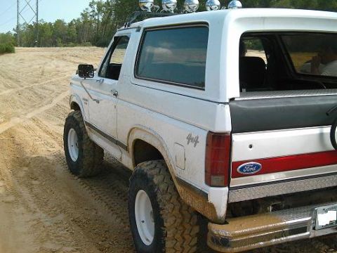 1986 Ford Bronco - A white Ford Bronco with off-road tires drives on a sandy track, with trees and a power line in the background.