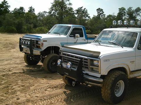 1986 Ford Bronco - Two lifted Ford pickup trucks are parked side-by-side on a sandy surface.