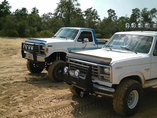 Two white Ford pickup trucks parked on sandy ground with trees in the background