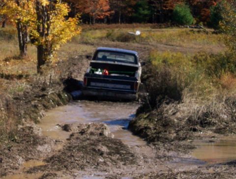 1979 Ford Bronco - A dark pickup truck navigates a muddy trail during autumn, with colorful trees in the background.