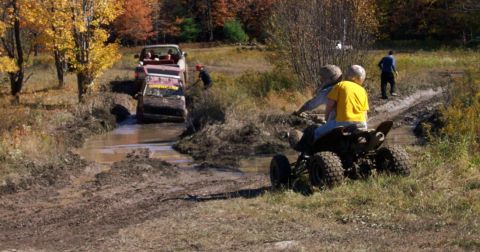 1979 Ford Bronco - ATV riders observe off-road vehicles navigating a challenging muddy terrain during an outdoor excursion.