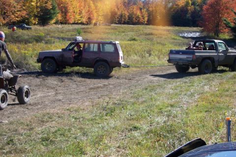 1979 Ford Bronco - A red SUV is being towed by a pickup truck across a muddy field during autumn.