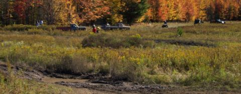 1979 Ford Bronco - Multiple vehicles navigate a muddy field during an off-road excursion with fall foliage visible.