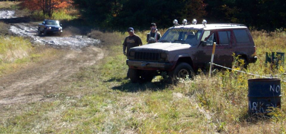 Maroon Jeep Cherokee XJ with roof lights on a muddy track, two people standing nearby