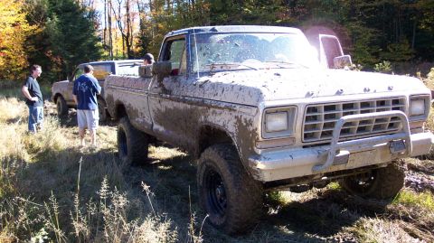 1979 Ford Bronco - A muddy pickup truck is stuck in the woods with two people observing the situation.