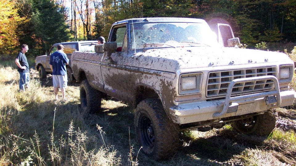 Muddy pickup truck with a brush guard, two people standing nearby, another vehicle behind it
