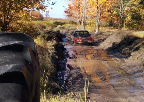 1979 Ford Bronco - A red pickup truck is stuck in a muddy path during autumn. A large tire is visible in the foreground.