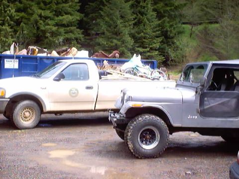1976 Ford F-100 - A silver Jeep CJ-7 is parked next to a white pickup truck, with a blue dumpster in the background.