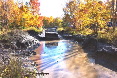 1979 Ford Bronco - A truck navigates a muddy trail surrounded by autumn trees, with another vehicle visible in the distance.