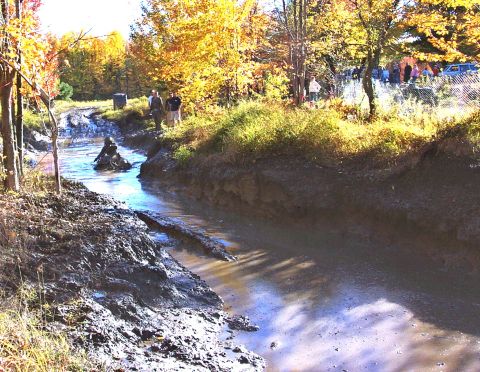 1979 Ford Bronco - An off-road vehicle splashes through a muddy watercourse during an event, with onlookers present.