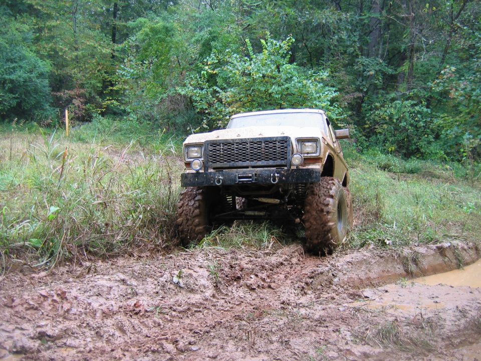 Lifted pickup truck with large tires driving through mud and water. Grille suggests late 1970s Ford