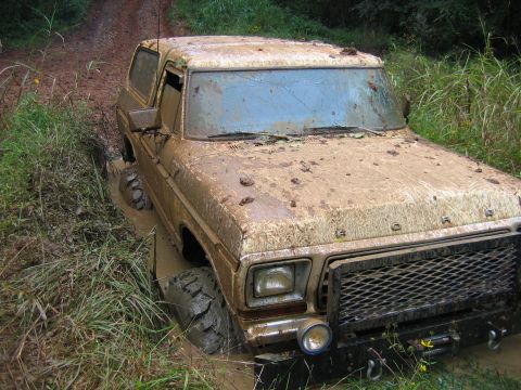 1992 Ford Mustang - This Ford Bronco is deeply stuck in mud on a dirt track, surrounded by vegetation.
