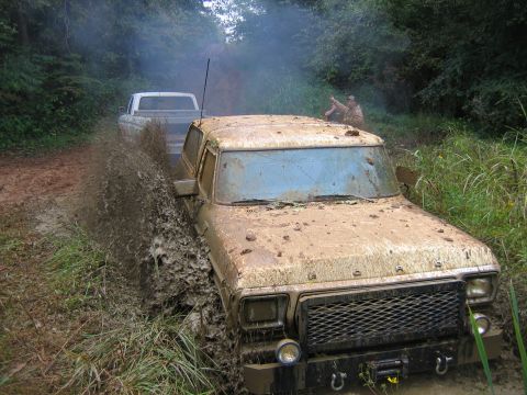 1992 Ford Mustang - Two trucks navigate a muddy off-road trail, with one getting stuck in deep mud.