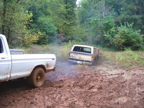 1992 Ford Mustang - A white pickup truck tows a stuck Ford Bronco out of a deep mud pit on a wooded trail.