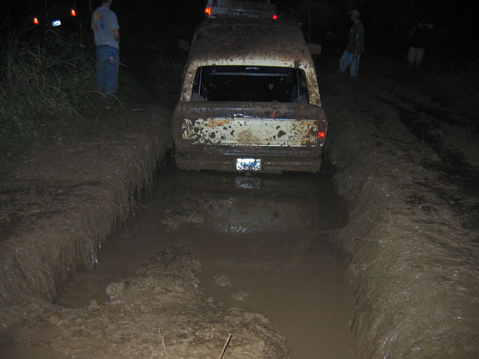 Rear view of a muddy white van stuck in a mud pit on a dirt track at night