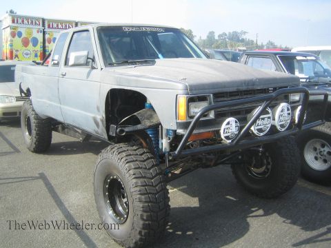 1990 Ford Bronco - gray primered lifted Chevrolet S-10 prerunner with a tubular front bumper, Hella lights and oversized tires sits in a parking lot near a ti…