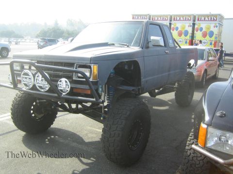 1990 Ford Bronco - blue-gray lifted mini-truck prerunner with a tubular front bumper, Hella lights and large mud tires parked beside other vehicles near a tic…