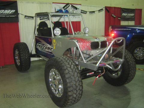 1990 Ford Bronco - silver tube-chassis rock buggy fitted with a BMW front clip and flame paint, wearing Pro Comp decals and mud tires, displayed at an indoor…