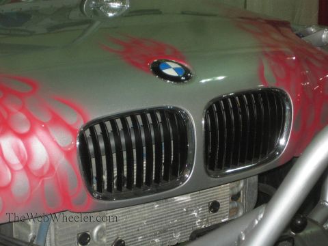 1990 Ford Bronco - close-up of a BMW kidney grille and blue-and-white roundel on a silver hood with red flame airbrushing, mounted on a tube-chassis vehicle.