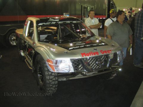 1990 Ford Bronco - silver Nissan trophy-style race truck with a roll cage and Nitto and Motive Gear decals on display at an indoor expo as spectators stand ne…