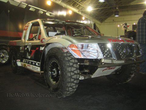 1990 Ford Bronco - silver Nissan Frontier prerunner race truck with a roll cage, Nitto decals and long-travel suspension displayed under lights at an indoor e…