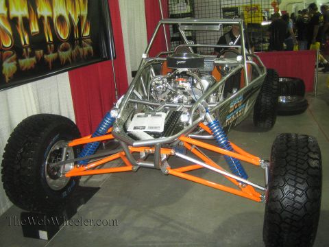 1990 Ford Bronco - tube-chassis sand rail buggy with an orange frame, blue coilover shocks and an exposed V8 engine sits at an indoor show booth with paddle t…