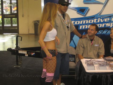1990 Ford Bronco - woman in a white top and fishnet stockings stands beside two men at a motorsports vendor booth with product displays at an indoor expo.