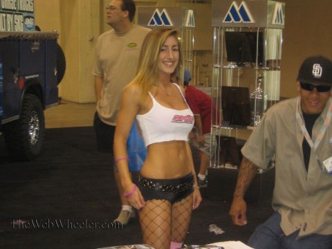 1990 Ford Bronco - woman in a white tank top and dark shorts walks past attendees and vendor displays at a busy indoor motorsports expo.